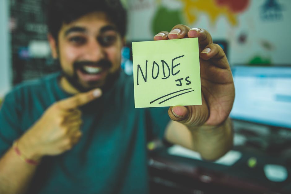 A cheerful software developer shows a Node.js sticky note in a tech workspace.